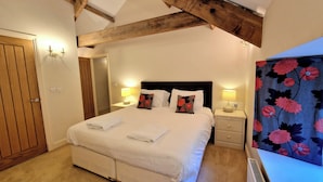 A bedroom with a large bed, wooden beams on the ceiling, two bedside tables with lamps, a door, and a radiator. - Clifton Cottage at Lovelady Shield (Alston)