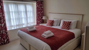 Een hotelkamer met een groot bed, wit beddengoed, rood-witte bloemen gordijnen, en een raam met dunne witte gordijnen.