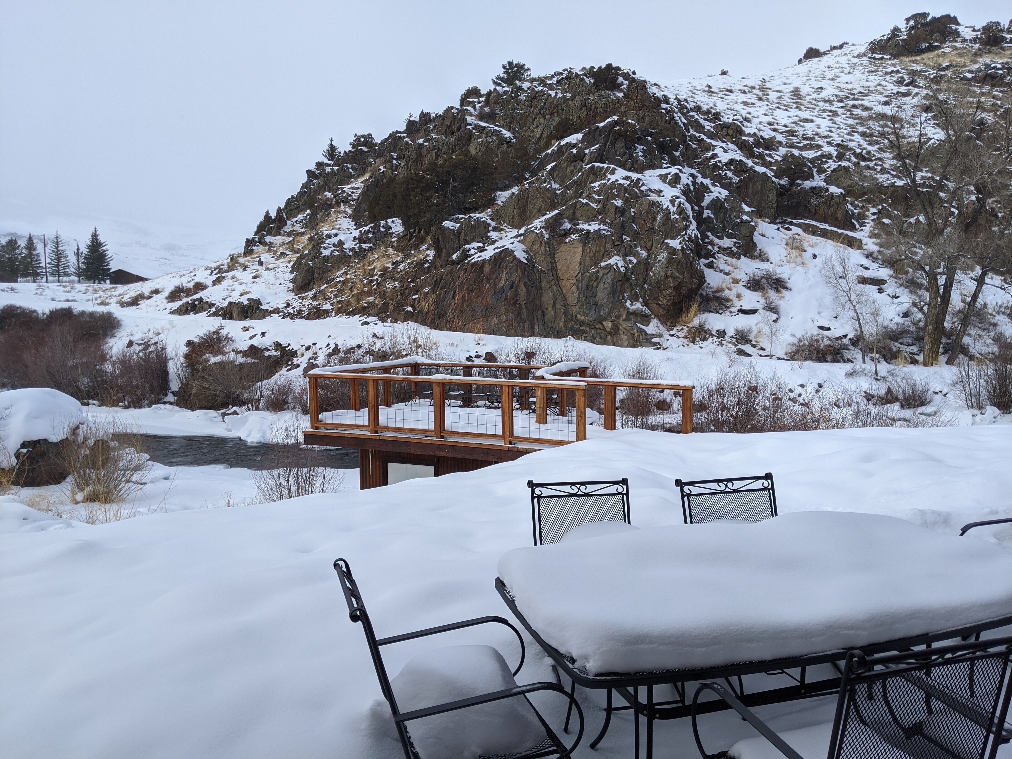 A snow-covered outdoor seating area with a table and chairs, overlooking a rocky mountain and a stream.
