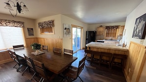 A dining area with a wooden table and chairs, a kitchen with wooden cabinets, and a large window with blinds. - Fabulous 5 BR Vacation Home on the Taylor River (Almont)