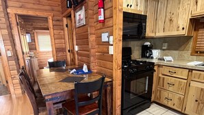 Interior de una cabaña de madera con una mesa de comedor, sillas, estufa y armarios de madera.