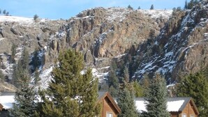 Una cabaña con fachada de madera, rodeada de árboles de hoja perenne y suelo cubierto de nieve, con una montaña rocosa al fondo.