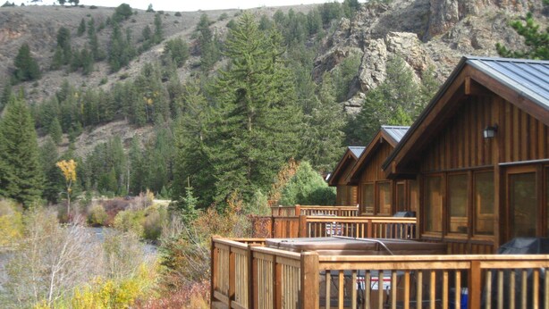A wooden cabin with a deck, surrounded by a forest and a rocky hillside.