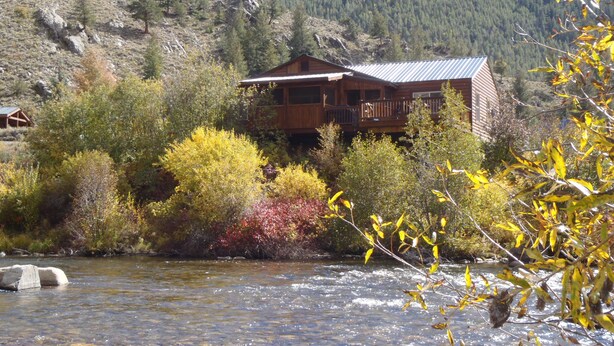 Eine Holzhütte mit Balkon, mit Blick auf einen Fluss, umgeben von Herbstlaub und einer Bergkulisse.