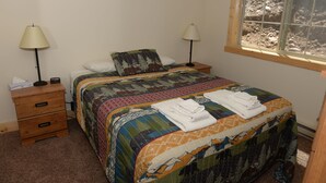 A neatly made bed with a colorful quilt, two bedside lamps, a wooden nightstand with drawers, and a window with a view of a rocky landscape. - Roomy Taylor Canyon Cabin With Private Fishing Access (Almont)