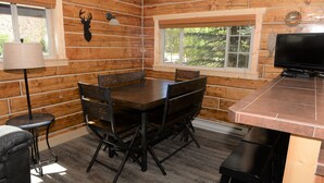 A wooden cabin interior with a dining table, chairs, a lamp, a television, and a fish wall decoration. - Roomy Taylor Canyon Cabin With Private Fishing Access (Almont)