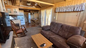 A cozy living area with a brown sofa, wooden coffee table, and a dining table with chairs. A kitchen with appliances and a dining area with a window are visible. - Modern "Tiny Home" Style Cabin on Taylor River with Private Fishing Access (Almont)