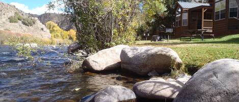 Una cabaña frente a un río, rodeada de árboles y con montañas al fondo.