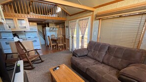 A cozy living room with a brown sofa, wooden coffee table, and a rocking chair. A kitchen area with cabinets and a refrigerator is visible in the background. - Modern "Tiny Home" Style Cabin on Taylor River with Private Fishing Access (Almont)