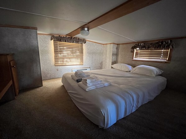 A bedroom with a large bed, two windows with blinds, a ceiling fan, and a wooden cabinet.