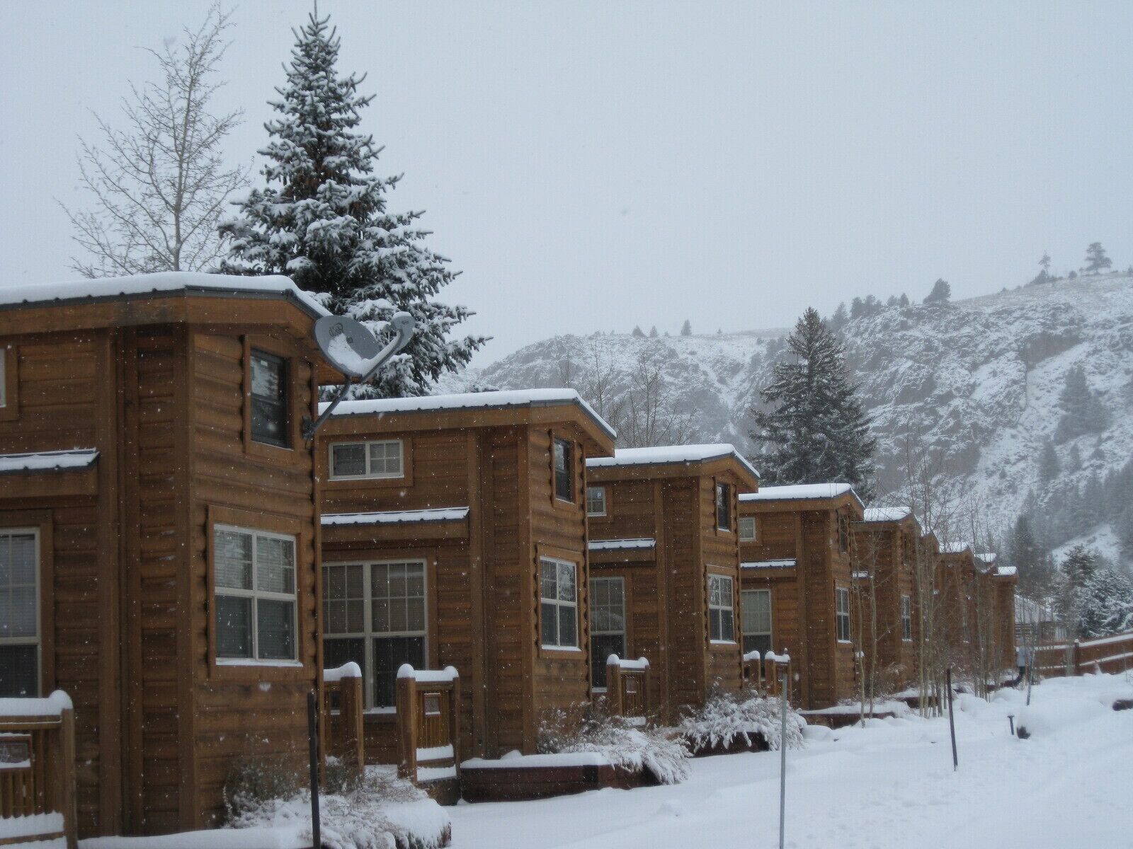 A row of wooden cabins with snow-covered roofs and windows, set against a snowy mountainous backdrop.