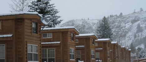 A row of wooden cabins with snow-covered roofs and windows, set against a snowy mountainous backdrop.