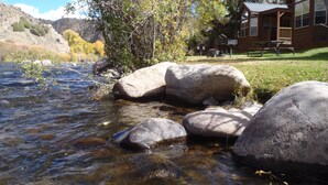 A cabin with a river in front, surrounded by trees and a mountainous backdrop.
