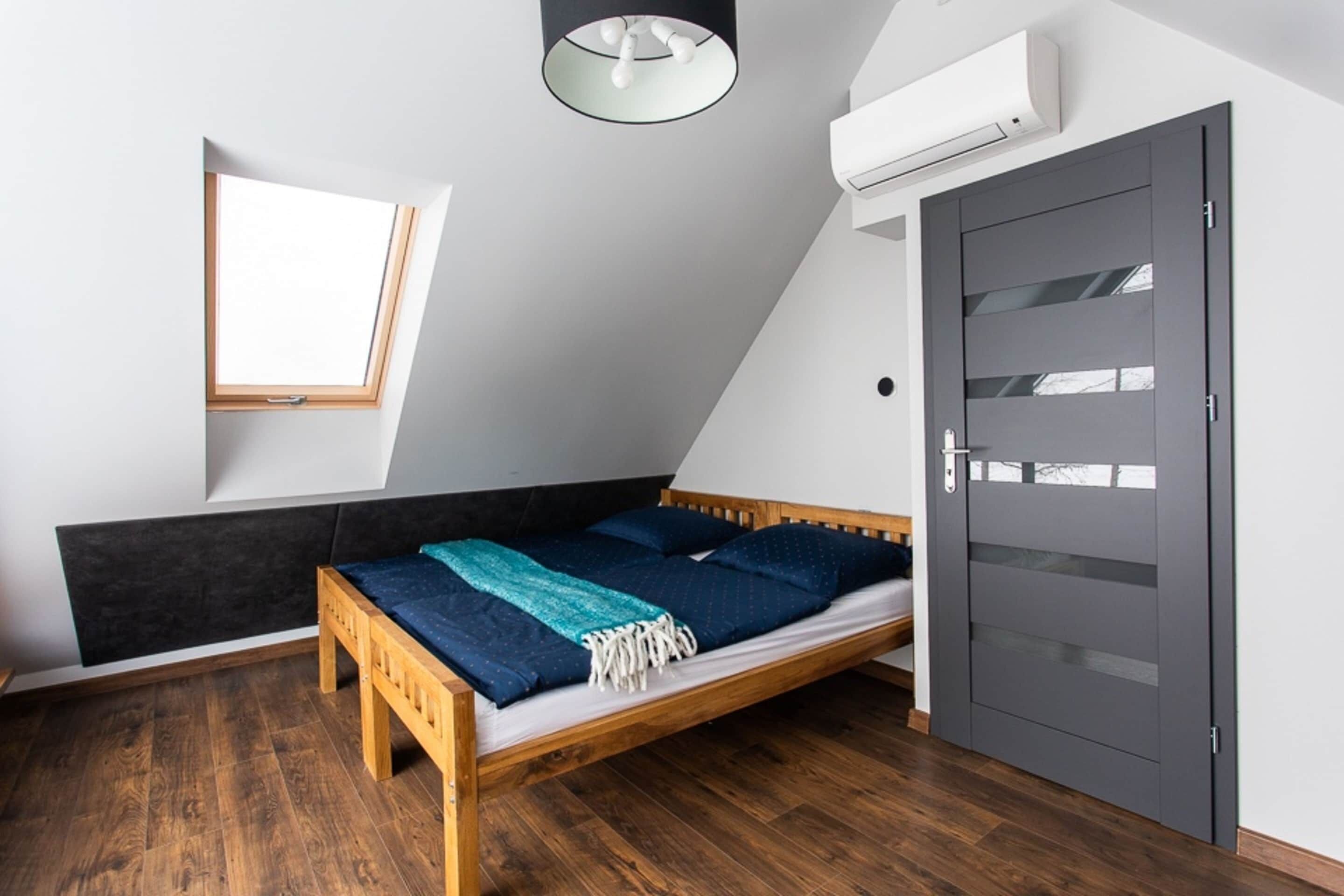 Camera da letto con soffitto spiovente, un lucernario, un letto con struttura in legno, una testiera scura, una porta grigia e un condizionatore d'aria bianco.