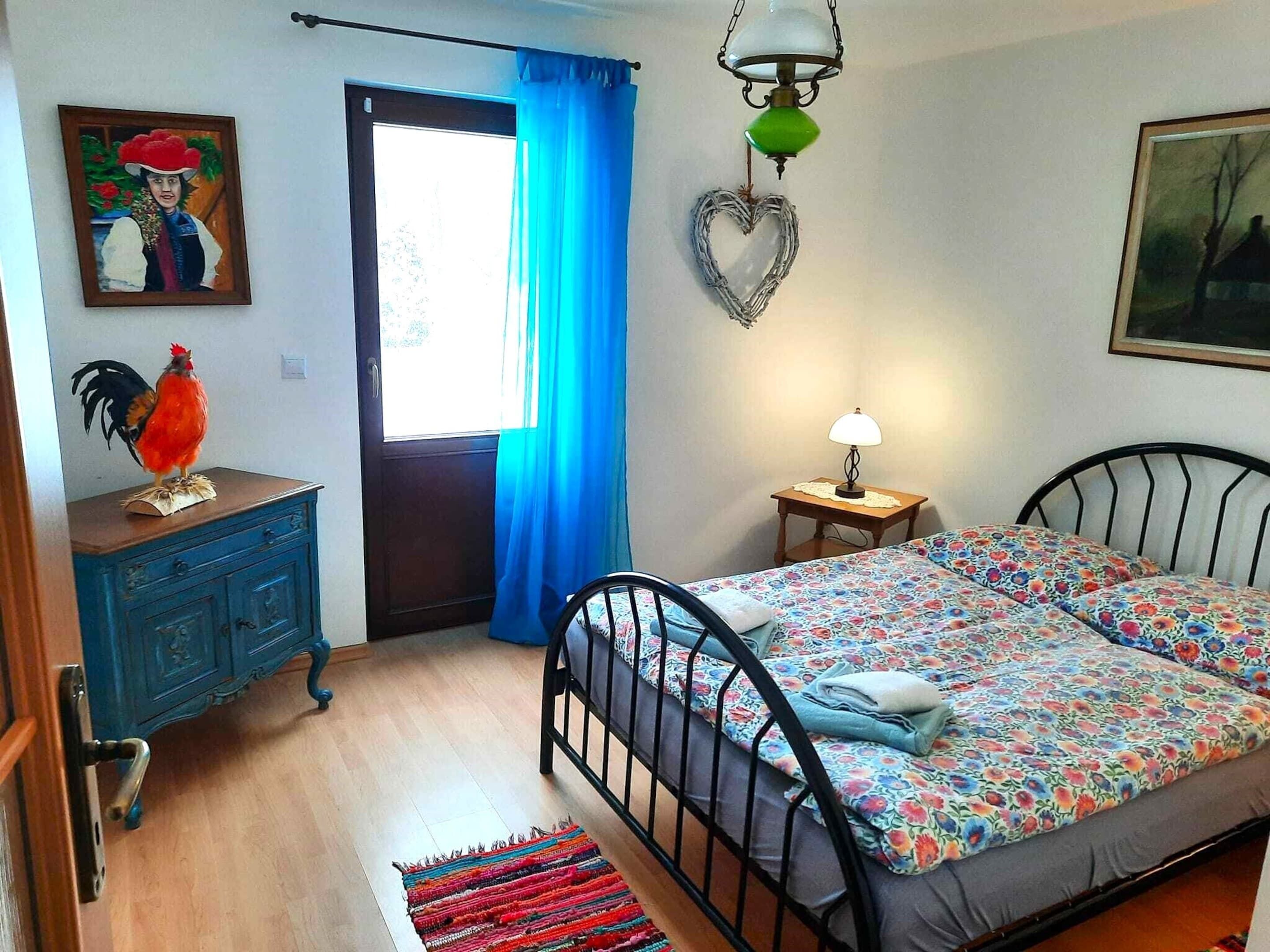 A bedroom with a bed, a dresser, a lamp, a heart-shaped decoration, and a painting on the wall.
