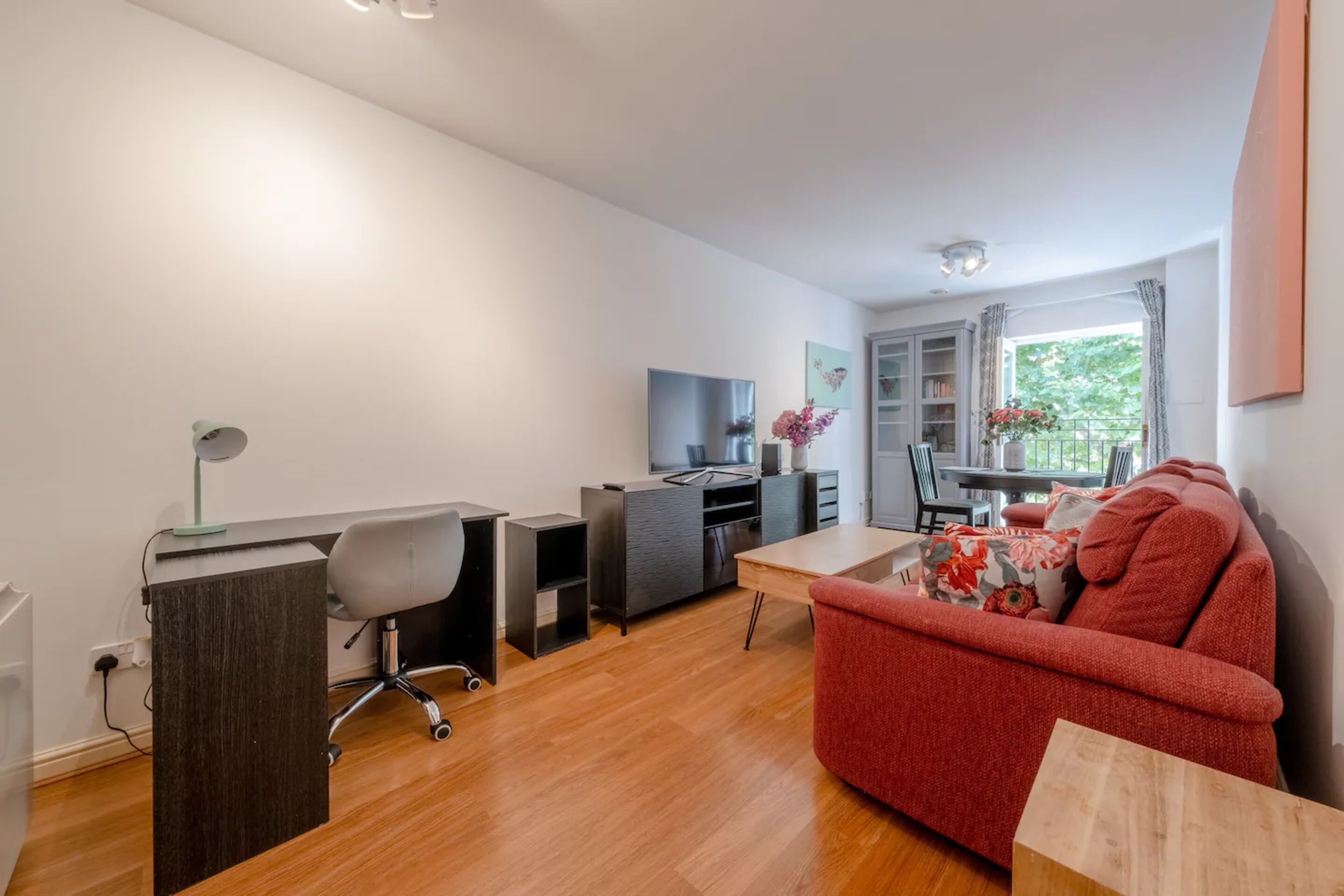Een moderne woonkamer met een rode bank, een houten salontafel, een bureau met stoel, een televisie en een boekenkast.