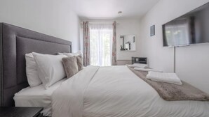 Ein ordentlich eingerichtetes Schlafzimmer mit einem großen Bett, einem Nachttisch, einem an der Wand montierten Flachbildfernseher und einem Fenster mit Vorhängen.