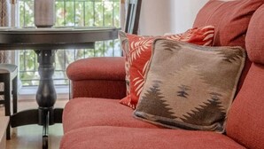 Ein rotes Sofa mit gemusterten Kissen, ein kleiner Tisch mit Blumen in einer Vase und Blick auf Grün hinter einem mit Vorhängen versehenen Fenster.
