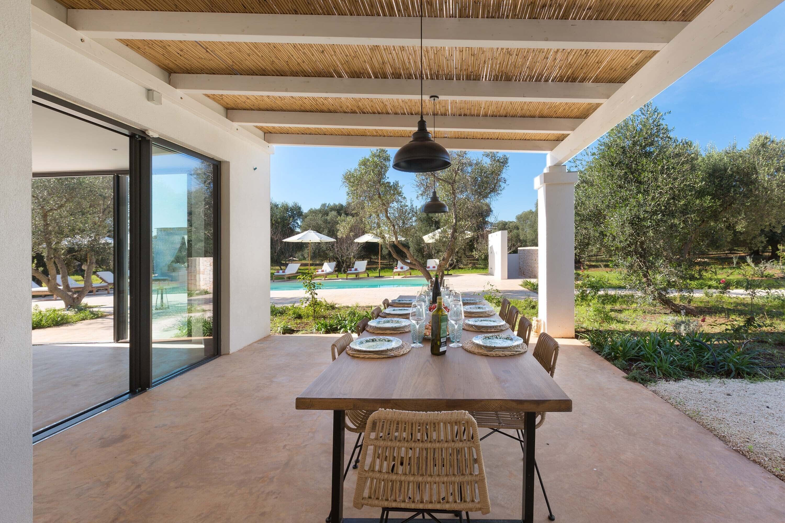 Een terras met een houten tafel voor zes personen, omringd door rieten stoelen, onder een pergola met een hanglamp.