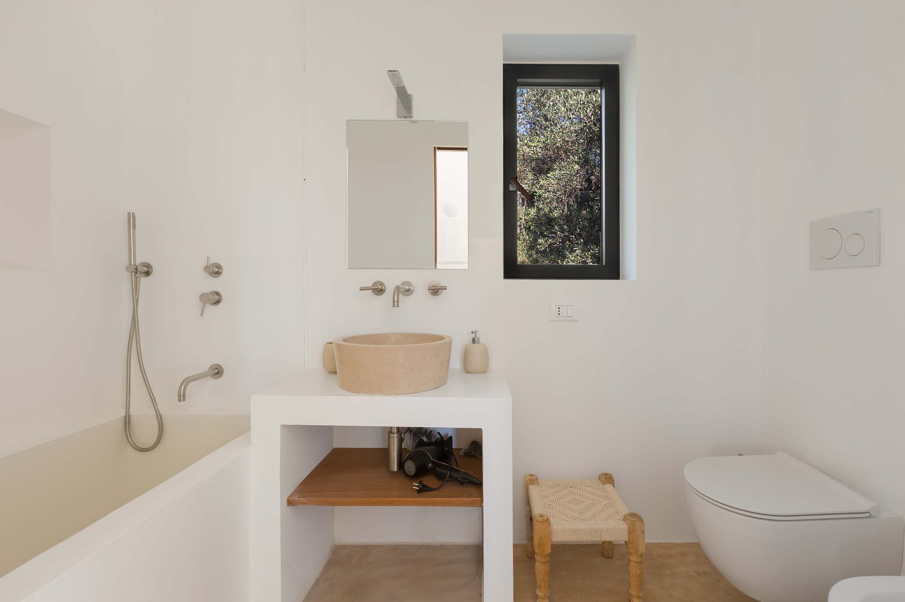 A modern bathroom with a bathtub, a sink with a mirror, a window with a view of trees, and a toilet.
