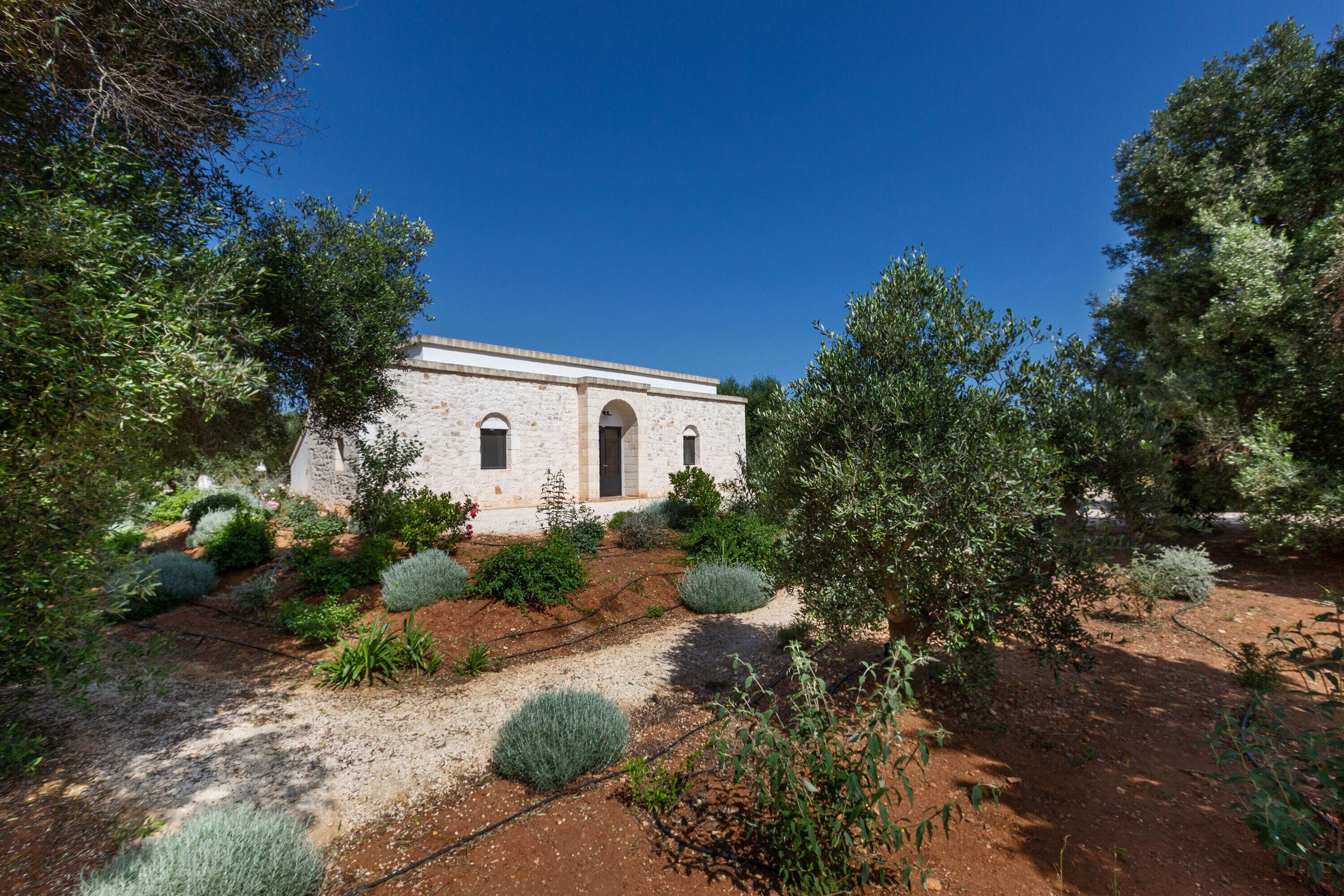 A white building with a central arched window and a door, surrounded by a garden with various shrubs and trees.