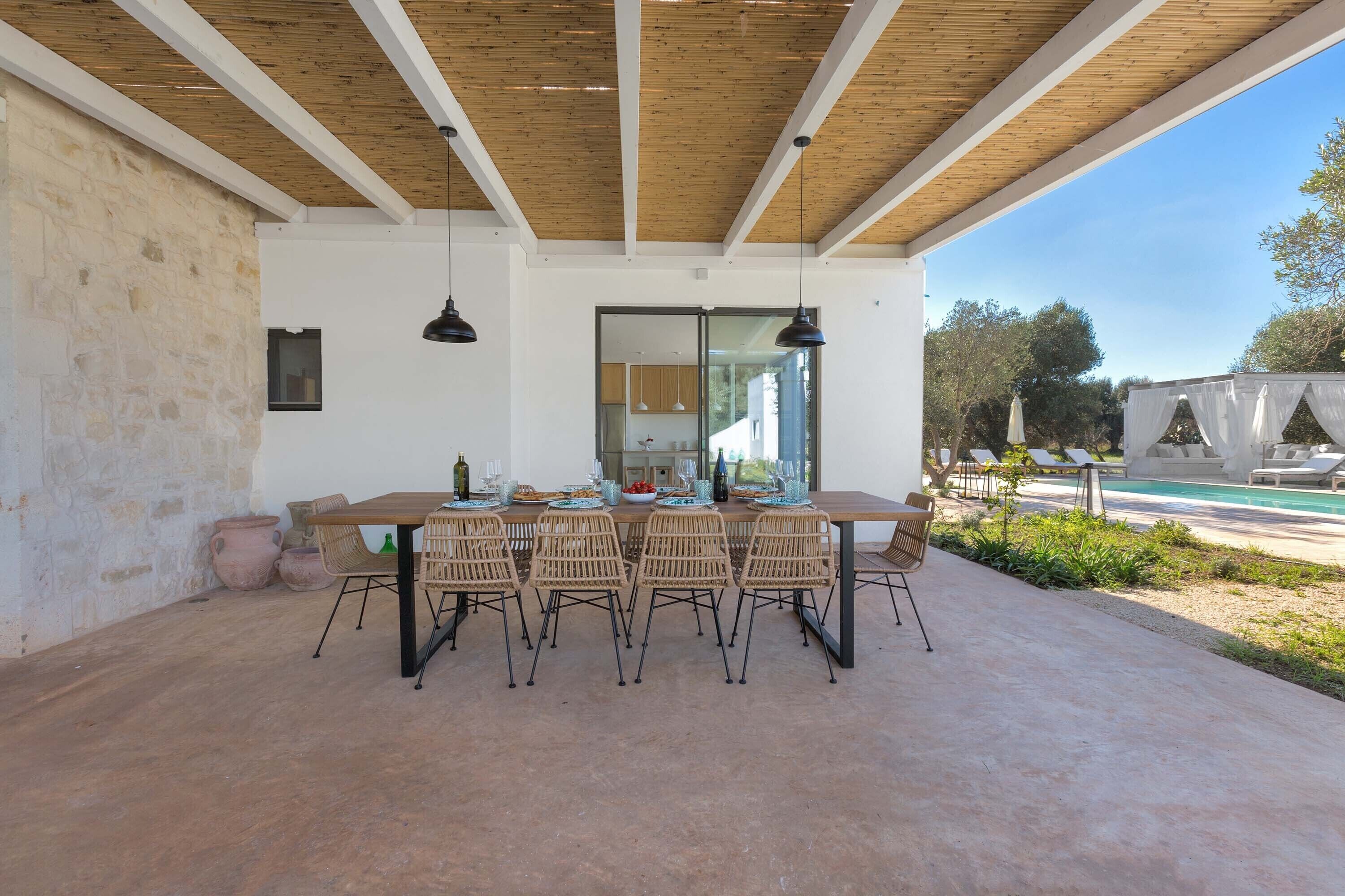 Een terras met eettafel, een houten plafond, een stenen muur en uitzicht op een zwembad en groen.