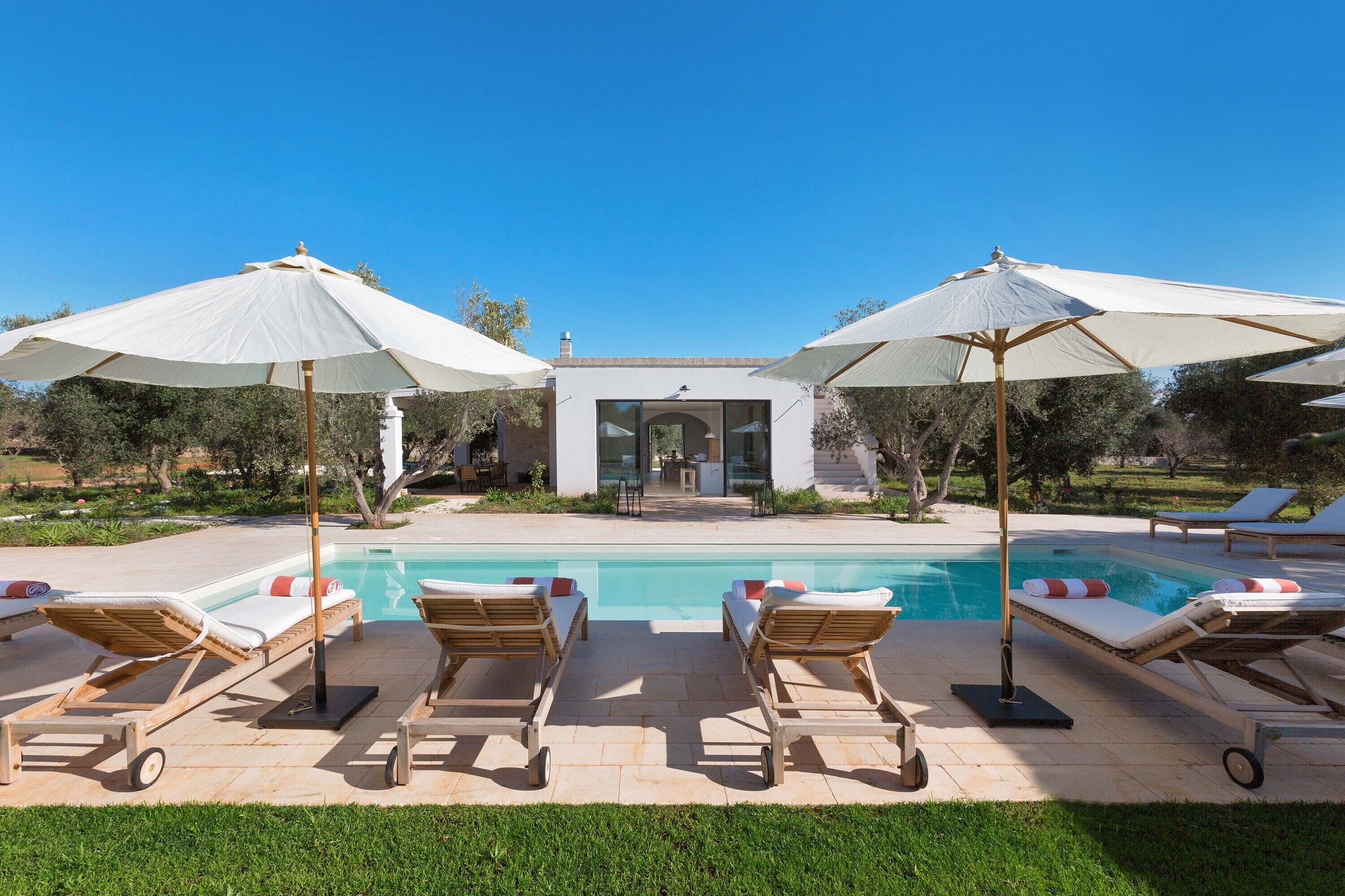 A pool area with lounge chairs and umbrellas, a clear sky, and a building in the background.