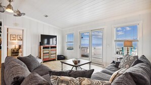 A spacious living room with a large gray sectional sofa, a small coffee table, and a flat-screen TV. There is a sliding glass door leading to a balcony with a view of the ocean. - Welcome to One Fish Two Fish! (St. George Island)