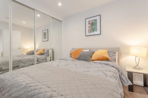 A bedroom with a large bed, a bedside table with a lamp, a mirror, and a framed picture on the wall. - Modern Flat in the Heart of West London (London)
