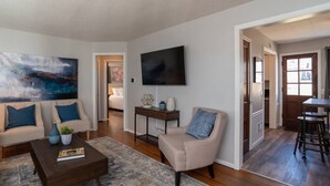 A living room with a sofa, coffee table, and a painting. There is a TV mounted on the wall and a door leading to another room. - Floyd's Retreat Stylish Home | Walk to Downtown Overland Park (Overland Park)