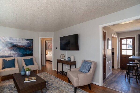 A living room with a sofa, coffee table, and a painting. There is a TV mounted on the wall and a door leading to another room.