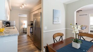 Una cocina moderna con un refrigerador de acero inoxidable, un comedor con una mesa y sillas de madera, y una sala de estar con un sofá color beige y un jarrón de orquídeas blancas.