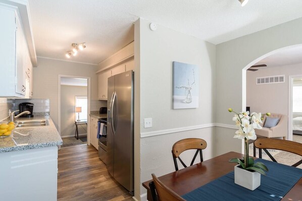 A modern kitchen with a stainless steel refrigerator, a dining area with a wooden table and chairs, and a living room with a beige sofa and a vase of white orchids.