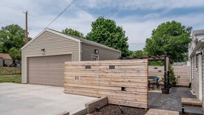 Ein Wohngrundstück mit Garage, Holzzaun, einer Terrassenfläche mit Tisch und Stühlen sowie einem kleinen Blumenbeet.