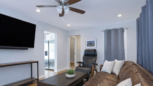 Ein modernes Wohnzimmer mit einem braunen Ledersofa, einem Holztisch, einem schwarzen Lederstuhl, einem Deckenventilator und einem flachbildfernseher an der Wand.