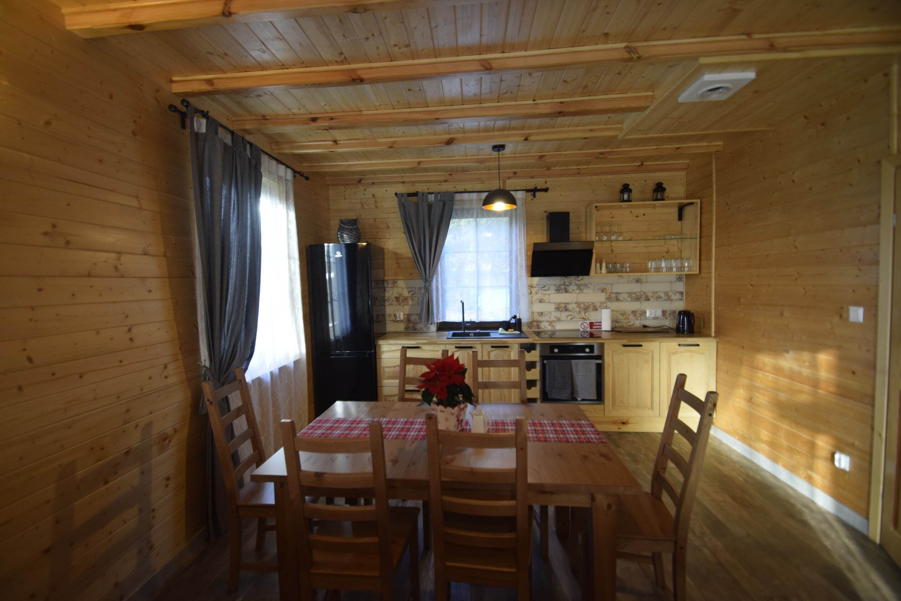 A wooden cabin interior with a dining table, chairs, a kitchen area with a refrigerator, stove, and shelves.