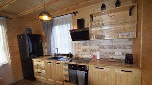 Cocina de cabaña de madera con un refrigerador negro, armarios de madera, una repisa de vidrio con vasos y una lámpara colgante.