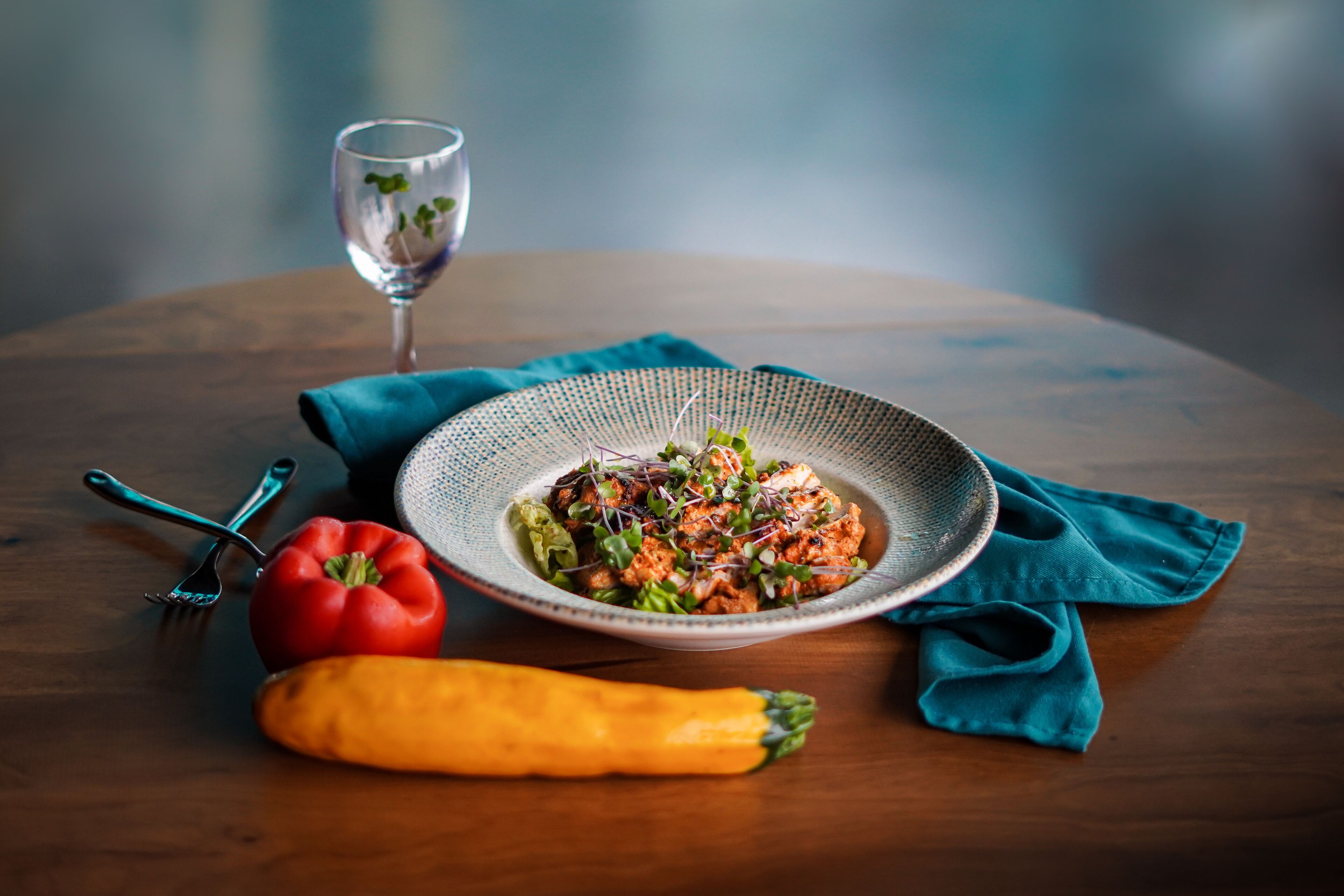 A round wooden table with a bowl of salad, a carrot, a bell pepper, and a glass of water.
