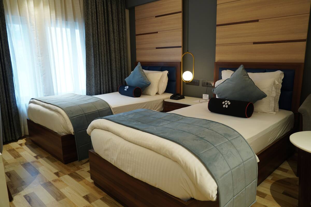 A hotel room with two beds, wooden headboards, a bedside table with a lamp, and a large window with curtains.