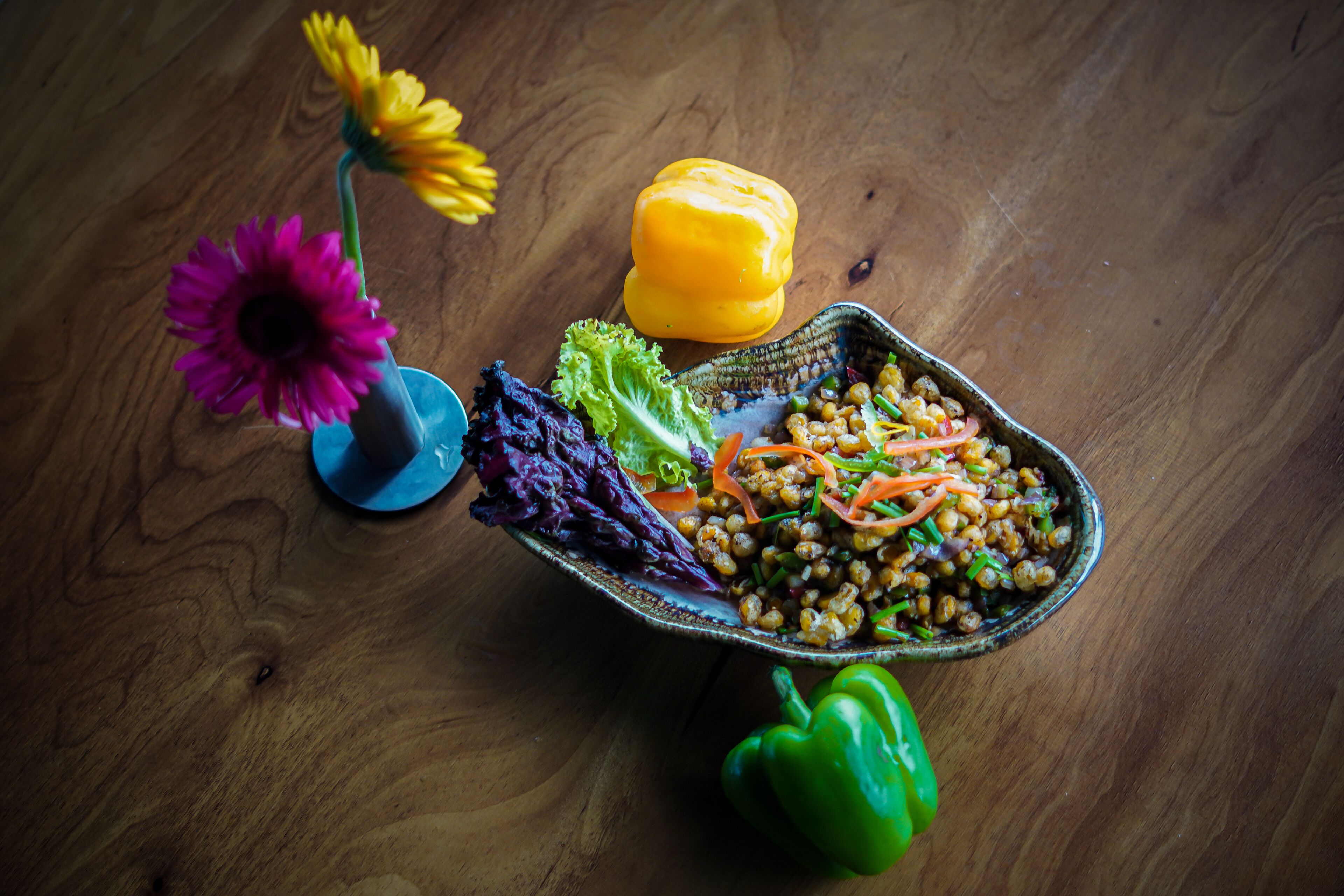 A wooden table with a bowl of salad, a yellow bell pepper, a green bell pepper, and a vase with flowers.