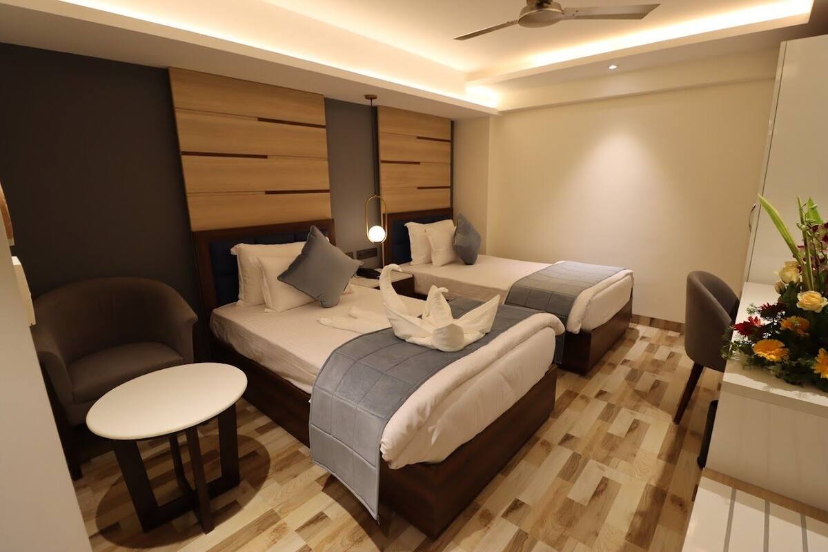 A modern hotel room with two beds, a small round table, a chair, and a desk with a vase of flowers.