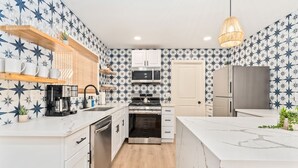 A modern kitchen with white cabinets, a built-in oven, microwave, and a refrigerator. There are wooden shelves with cups and a hanging light fixture. - New Listing! Desert Oasis ~ Hot Tub (Twentynine Palms)