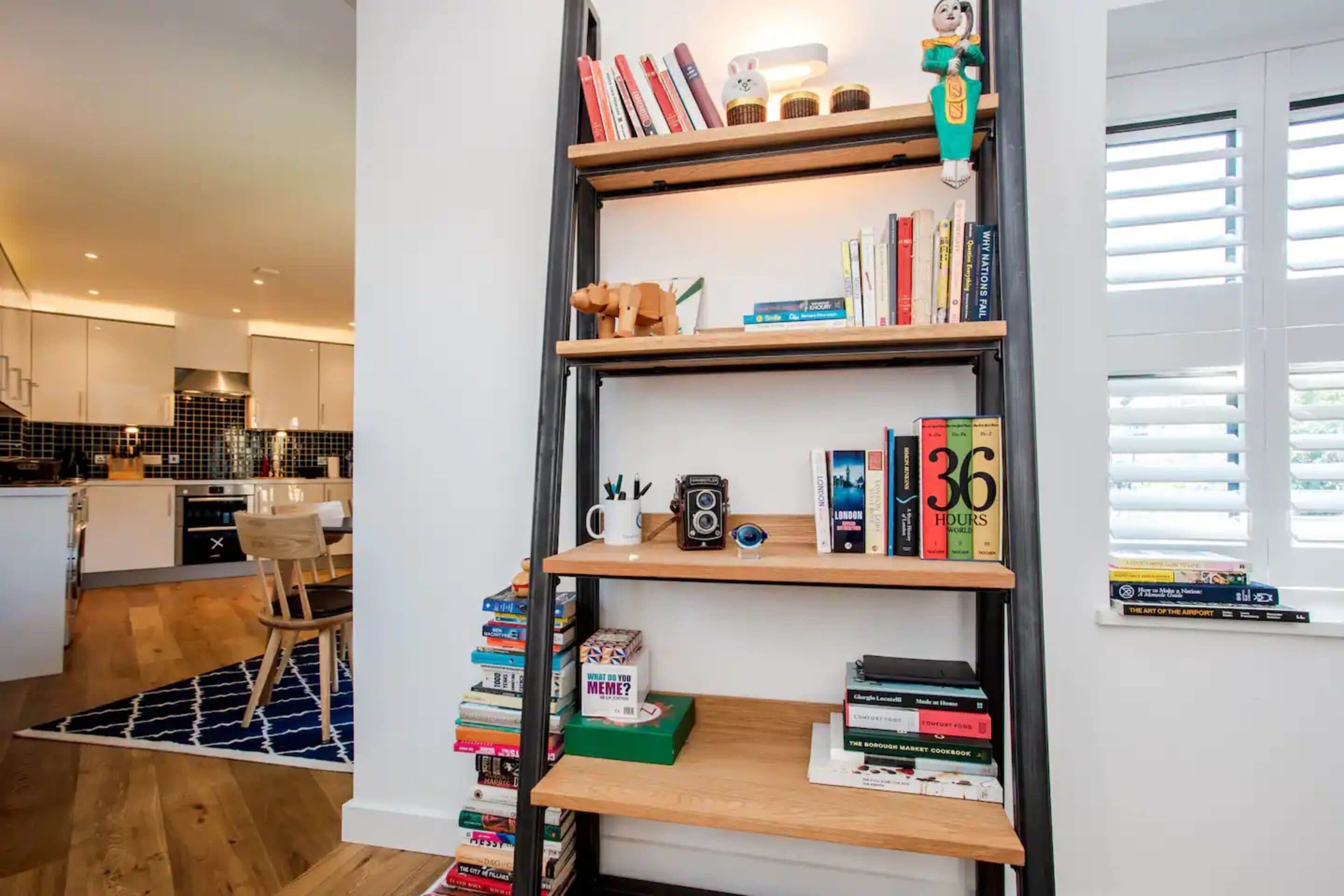 A modern kitchen with white cabinets, a wooden dining table, and a bookshelf filled with books and decorative items.