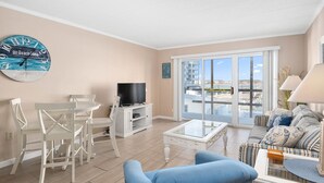A living room with a TV, white furniture, a blue sofa, and a large window with a view.