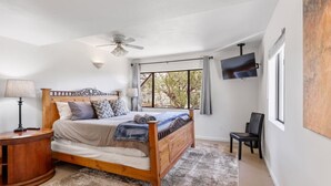 Ein Schlafzimmer mit einem Holzbett, einem Deckenventilator, einem an der Wand montierten Fernseher und einem Fenster mit Blick auf Bäume.