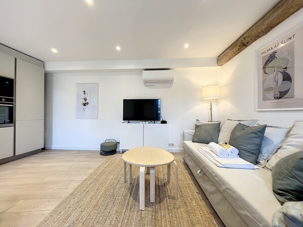 A modern living room with a sofa, a small round table, a flat-screen TV, and a kitchen area with cabinets and an oven.