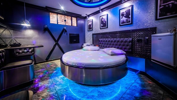 A modern bedroom with a circular bed, a vanity with a mirror, and a small refrigerator. - Motel Dallas (Cascavel)
