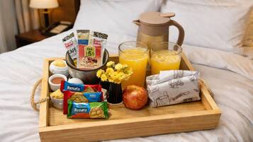 A breakfast tray on a bed with a coffee pot, juice glasses, snacks, and a small vase of flowers.