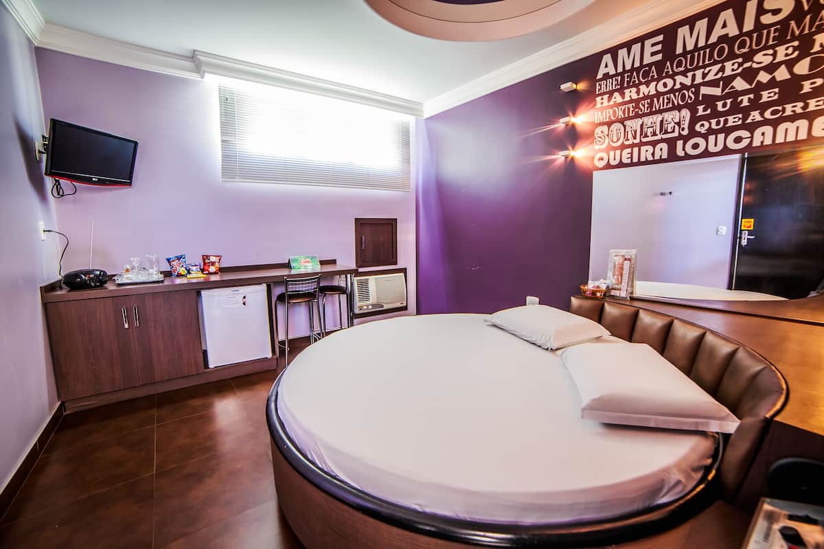 A hotel room with a round bed, a desk, a chair, a television, and a window.