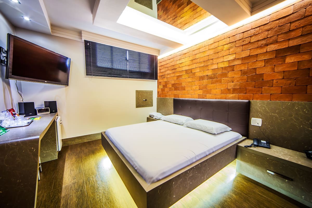 A modern hotel room with a flat-screen TV, a desk with a computer, a bed with a headboard, and a brick-patterned wall.